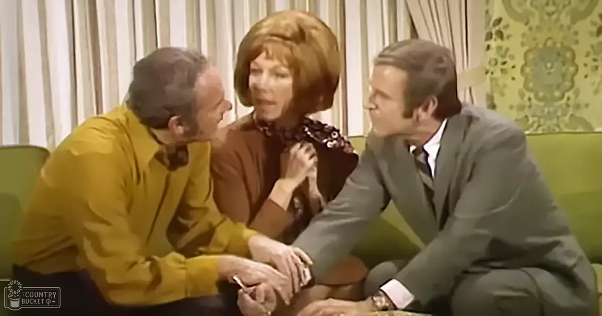 An adult male singer wearing a mustard-yellow long-sleeved shirt with a patterned tie or scarf, seated among other adults in a mid-20th century-style living room setting with green furniture and striped curtains.