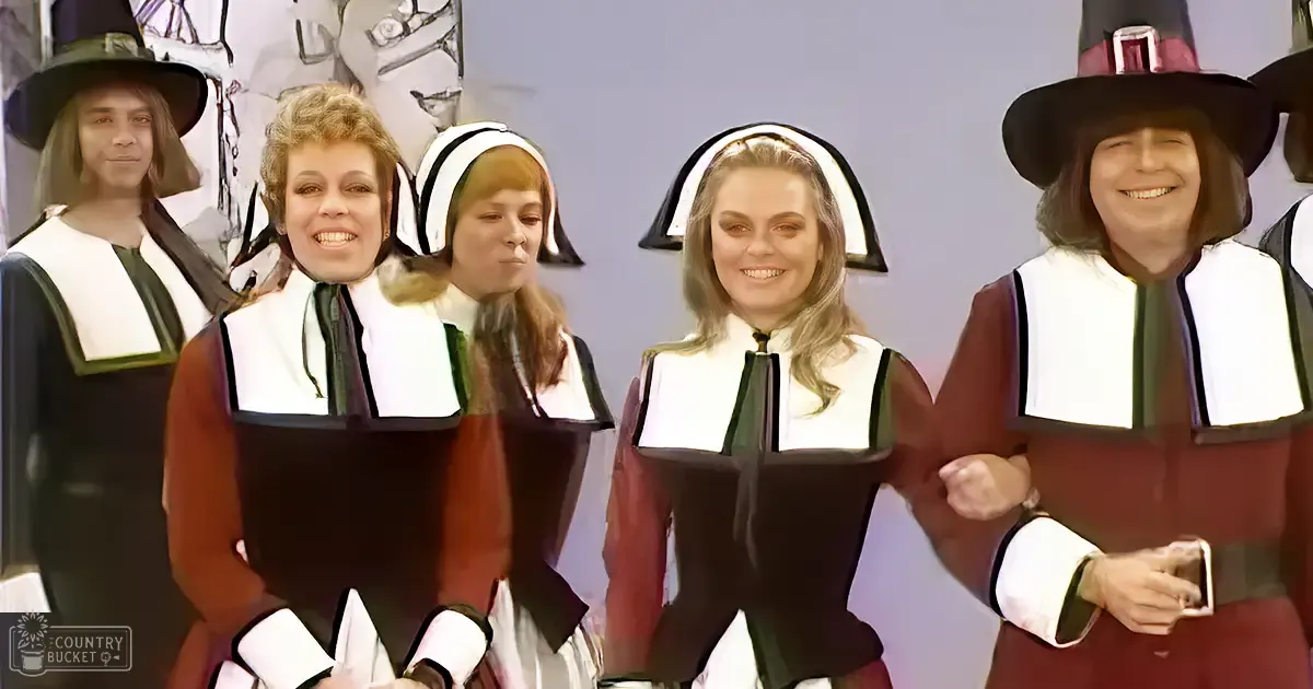 A group of individuals dressed in pilgrim costumes, possibly for a theatrical performance or themed event, indoors with red dresses, white collars, black hats, and beige background.