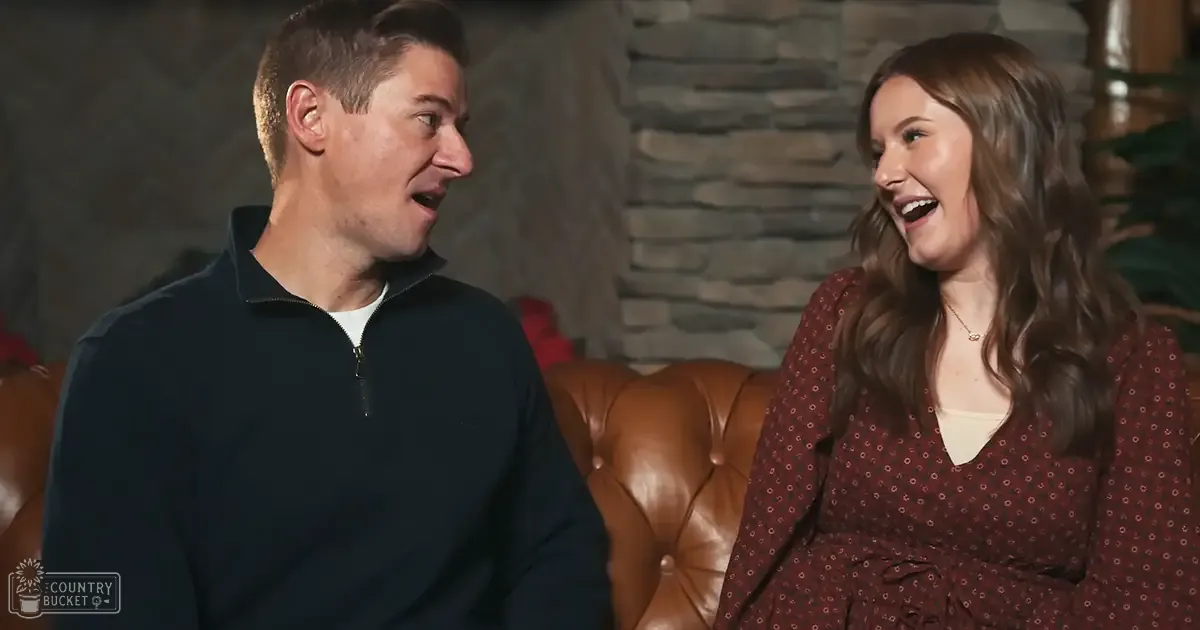 A couple indoors, male adult wearing a dark blue zip-up sweater over a white t-shirt and smiling at his female companion in a maroon dress with light patterns and a gold necklace pendant. A tan leather couch and stone wall backdrop suggest an indoor setting with cozy ambiance.
