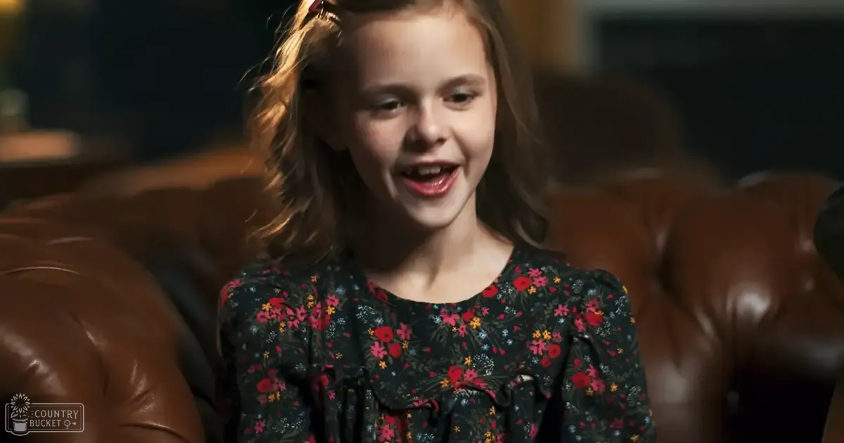 A young child wearing a dark floral dress with ruffled sleeves sits indoors in a living room, surrounded by furniture and home decor under warm lighting.