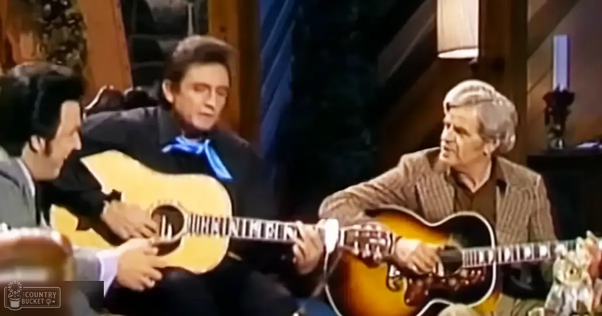 An adult male singer wearing a light-colored suit jacket and dress shirt performs alongside another adult in a dark, casual shirt with a blue bandana, both on an indoor stage with rustic wooden paneling and warm lighting.