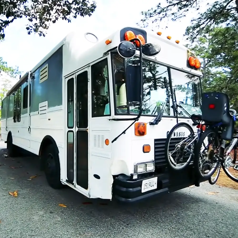 A white bus with a bicycle mounted on its front, located in an outdoor setting with trees visible in the background. The scene suggests spring or summer due to greenery and mild weather.