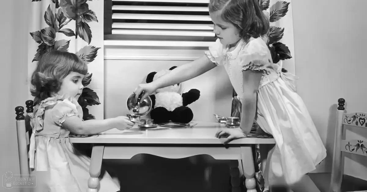 Two young female children playing tea party indoors with a stuffed panda bear, floral curtains, and natural daylight.