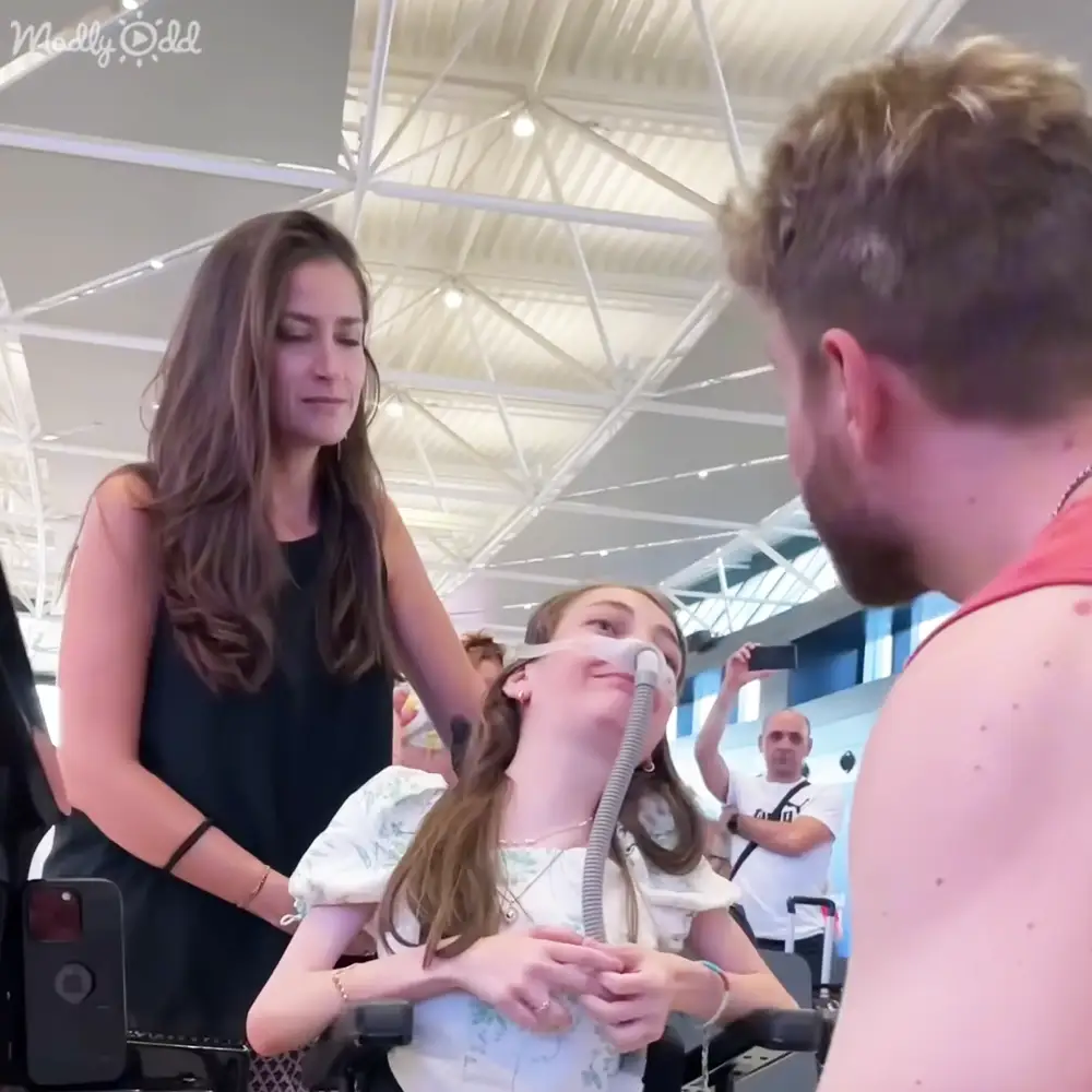 Young adult female in a white floral-patterned top and necklace, seated in a wheelchair with an oxygen mask at an airport terminal.