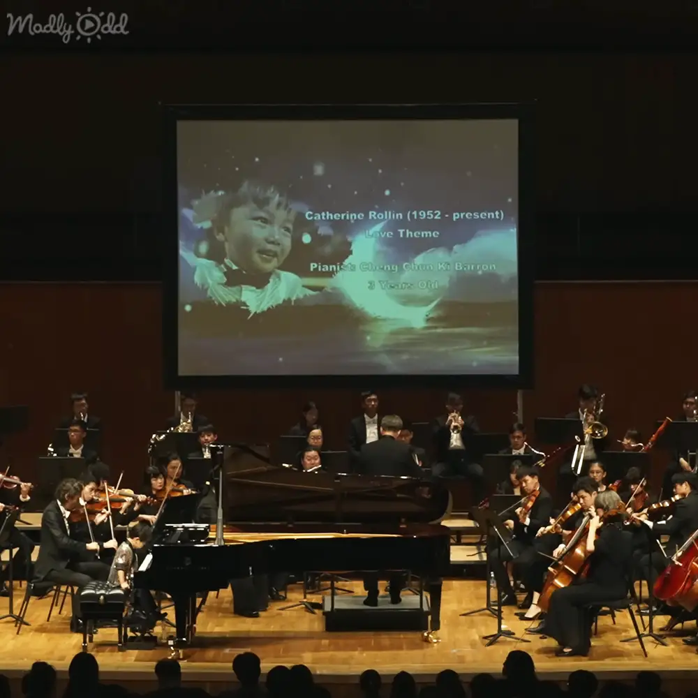 A young child playing piano at an indoor concert hall with musicians in the background.