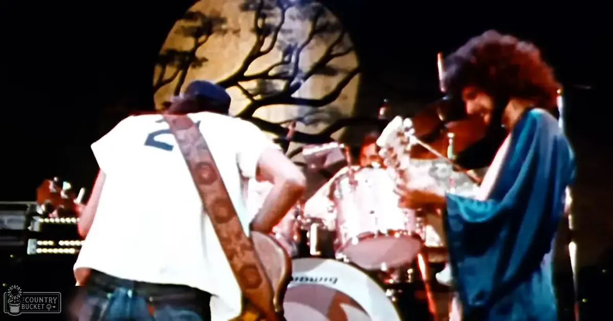 A live music performance at an outdoor concert venue with a young adult male singer in a white t-shirt and blue jeans, and another adult male wearing a patterned blue jacket or robe; drum set visible behind the performers under dark nighttime sky.