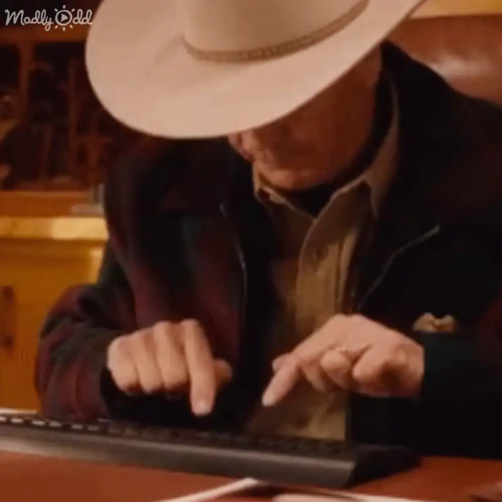 Older adult male wearing a wide-brimmed white hat with dark band, dark jacket, light brown shirt or sweater in an indoor office or workspace setting with natural daylight and wooden furniture.