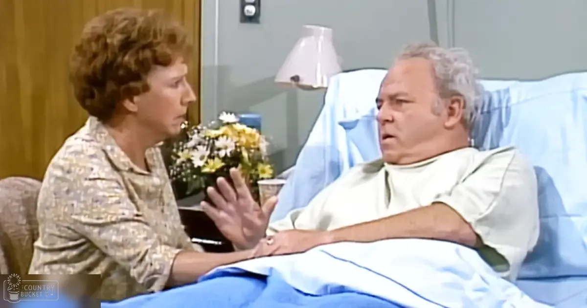 An older adult female in a patterned blouse and an older adult male in a hospital gown, surrounded by clinical furniture and a bouquet of yellow and white daisies in a hospital room with light-colored walls and wood paneling.