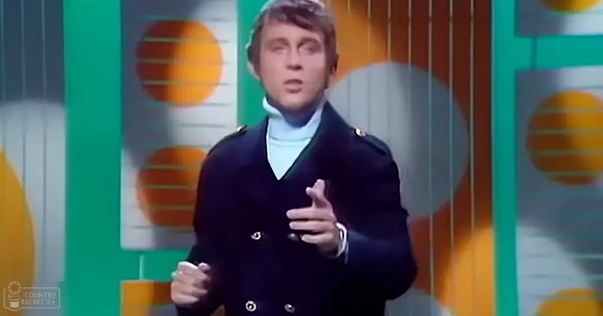 Adult male performer wearing a dark double-breasted coat with epaulets, standing against an orange and green backdrop. 'Madly Odd' watermark suggests studio setting for entertainment show in the 1970s.