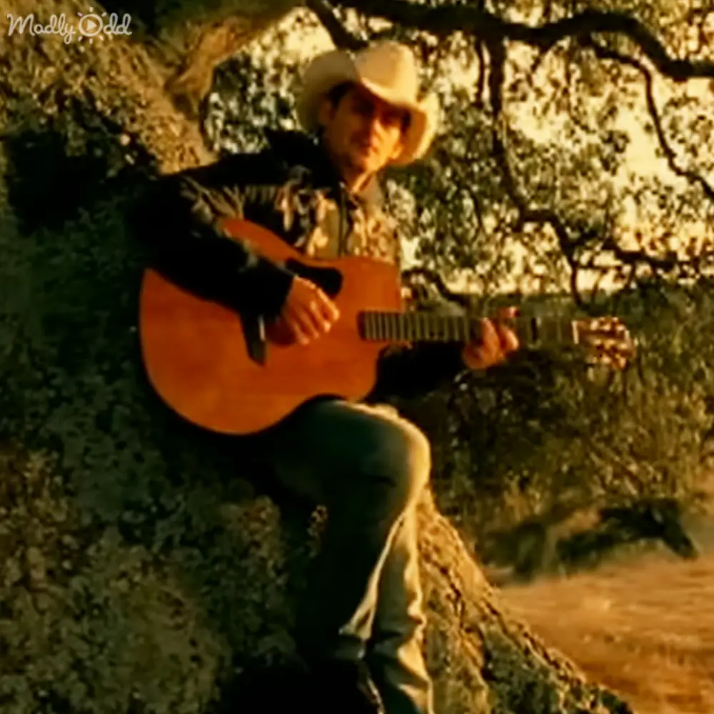 A young adult male plays guitar outdoors amidst a forested natural setting with warm sunlight filtering through trees, wearing a cowboy hat and patterned shirt.
