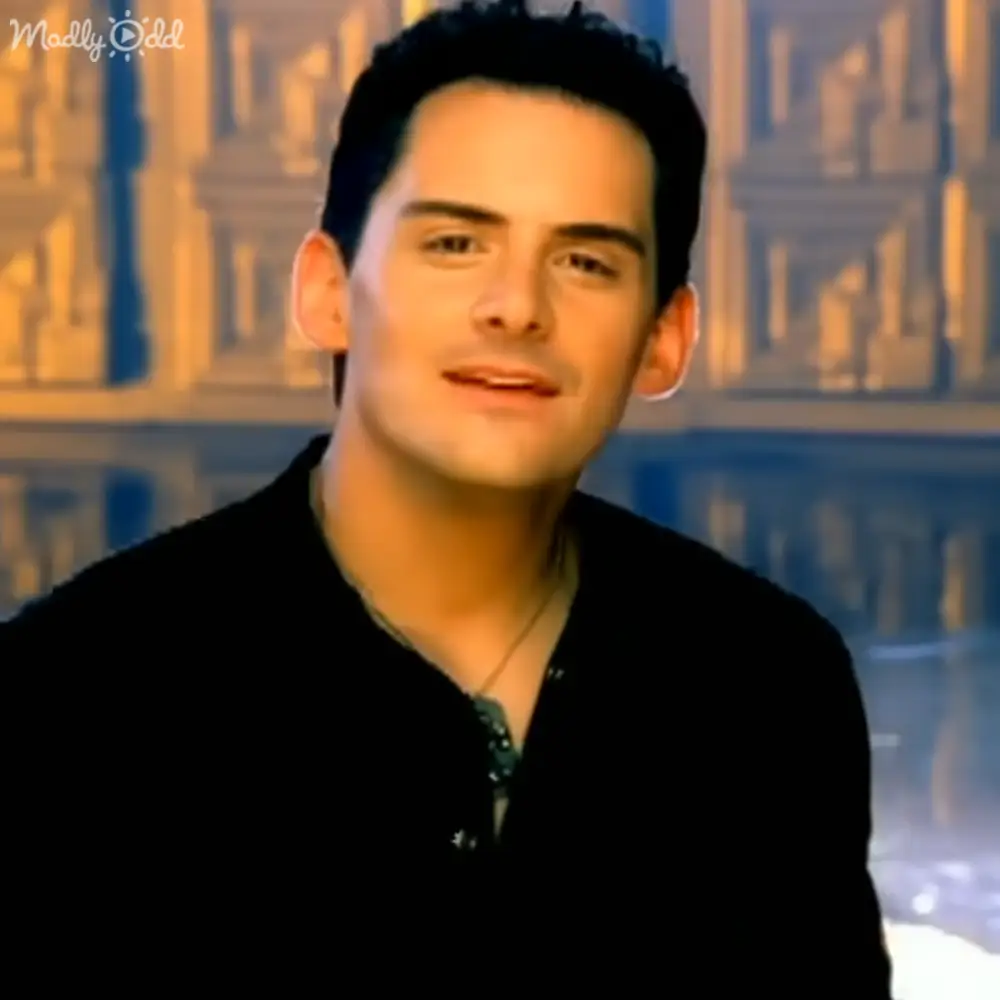 Young adult male wearing a black V-neck long-sleeved shirt indoors with 'Madly Odd' text in top left corner.