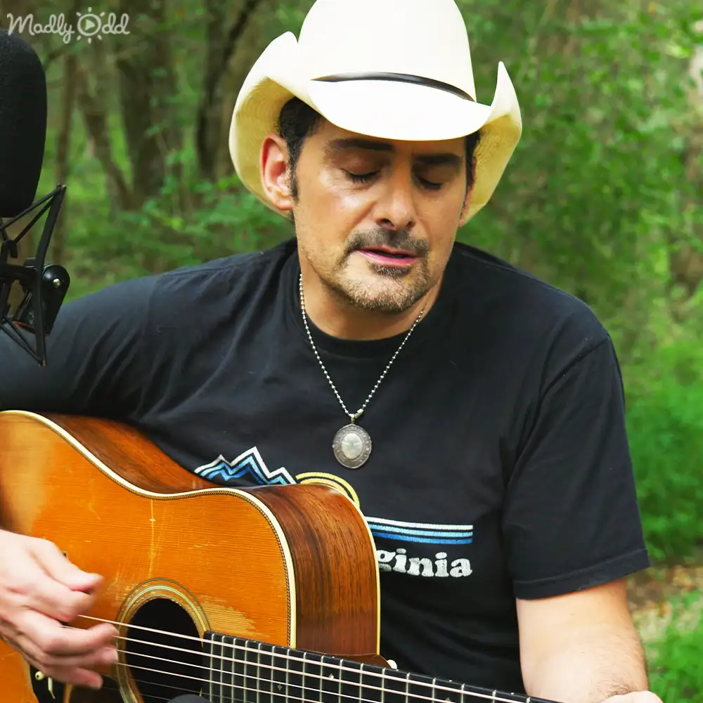 A middle-aged male singer in a casual black T-shirt with 'Virginia' text and graphic, playing an acoustic guitar outdoors during daylight.