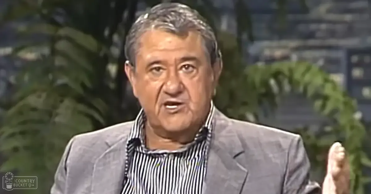 An older adult male in a studio with blurred greenery in the background, wearing a gray blazer over a striped button-up shirt.