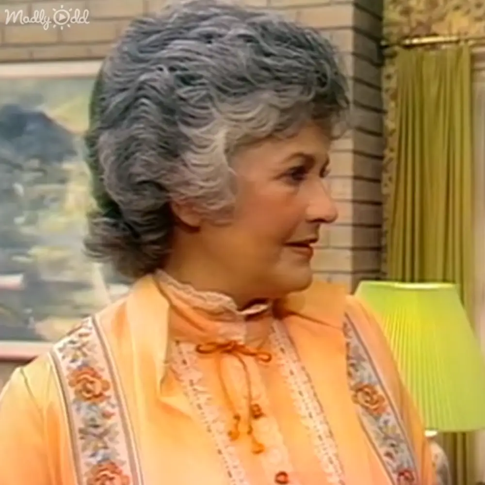 An older adult woman wearing a light orange floral blouse indoors with warm lighting, green lampshade visible in the background.
