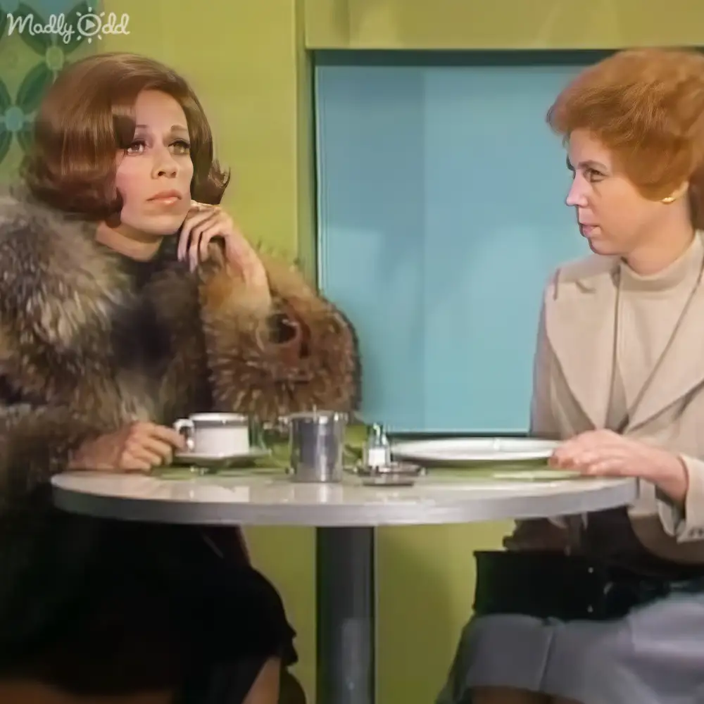 A middle-aged adult female and another person seated at a table in an indoor café setting with bright pastel walls; a cow is partially visible behind one of the individuals, adding an unexpected element to the scene.