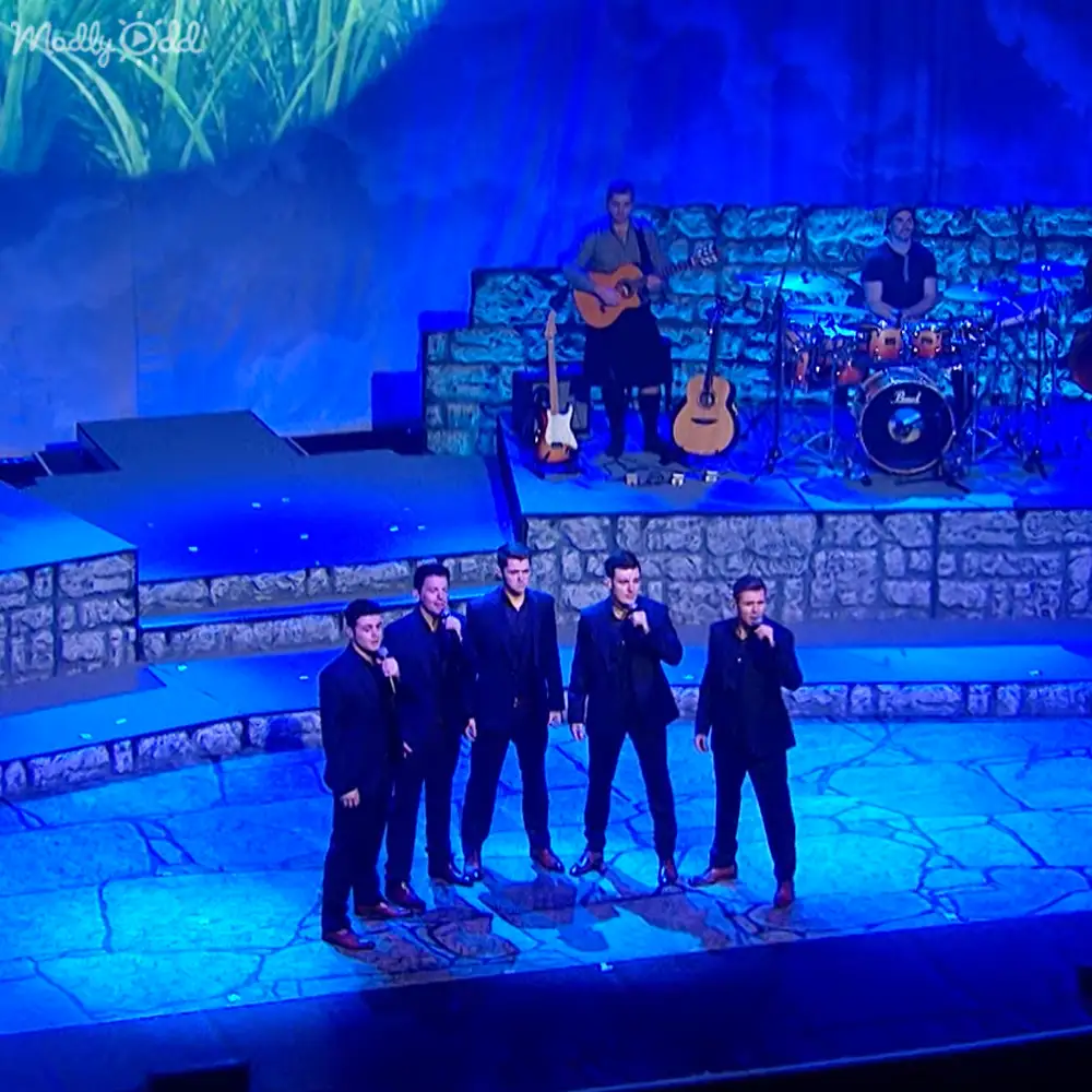 A group of adult male singers in formal attire performing on a stage with blue lighting effects at an indoor concert venue.