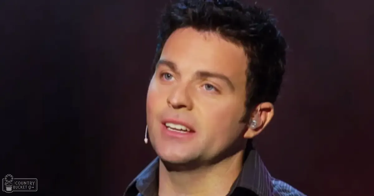 Young adult male wearing a dark blue button-up shirt indoors under dim lighting suggestive of a performance or interview setting.