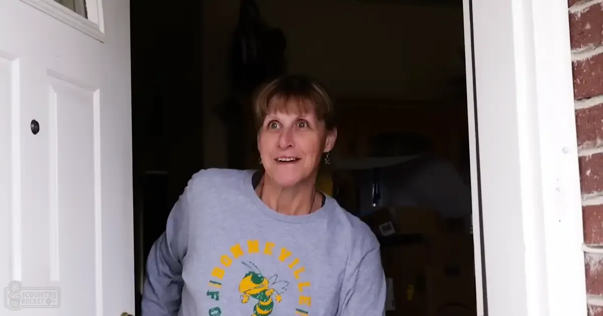 An older adult female wearing a light gray sweatshirt with 'Bonneville' text and bee graphic, in a residential setting with natural daylight.