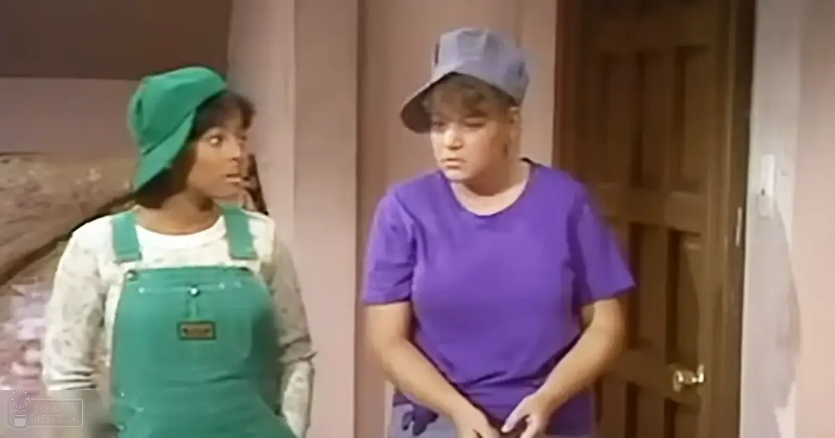 Two young adults, one in green overalls with a beret-style hat and another in a purple t-shirt with a gray baseball cap, indoors with wooden door and light-colored walls