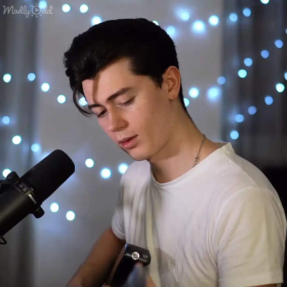 A young adult male in a relaxed white t-shirt, thin chain necklace, and indoor setting with string lights for ambiance.