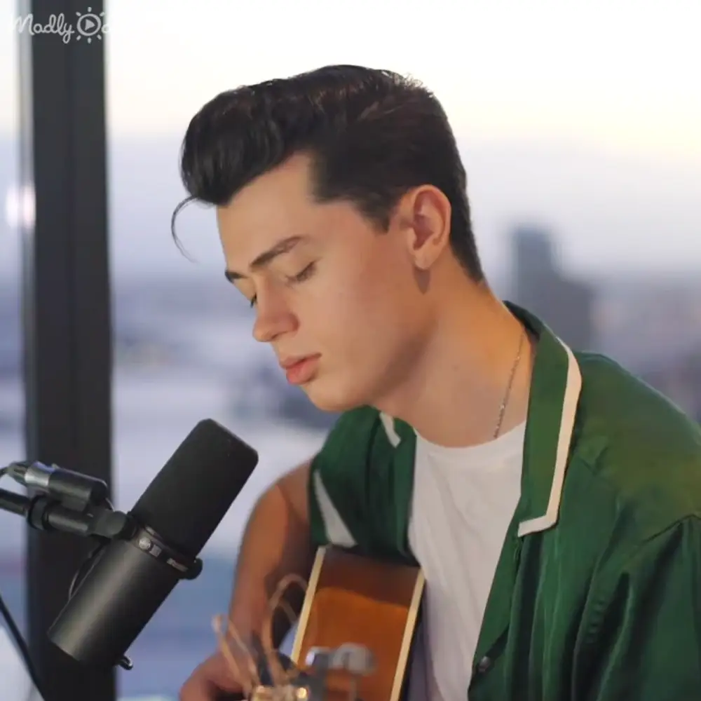 A young adult male wearing a white t-shirt under a green jacket with white trim is positioned indoors near large windows showing a cityscape, suggesting he might be recording or performing.