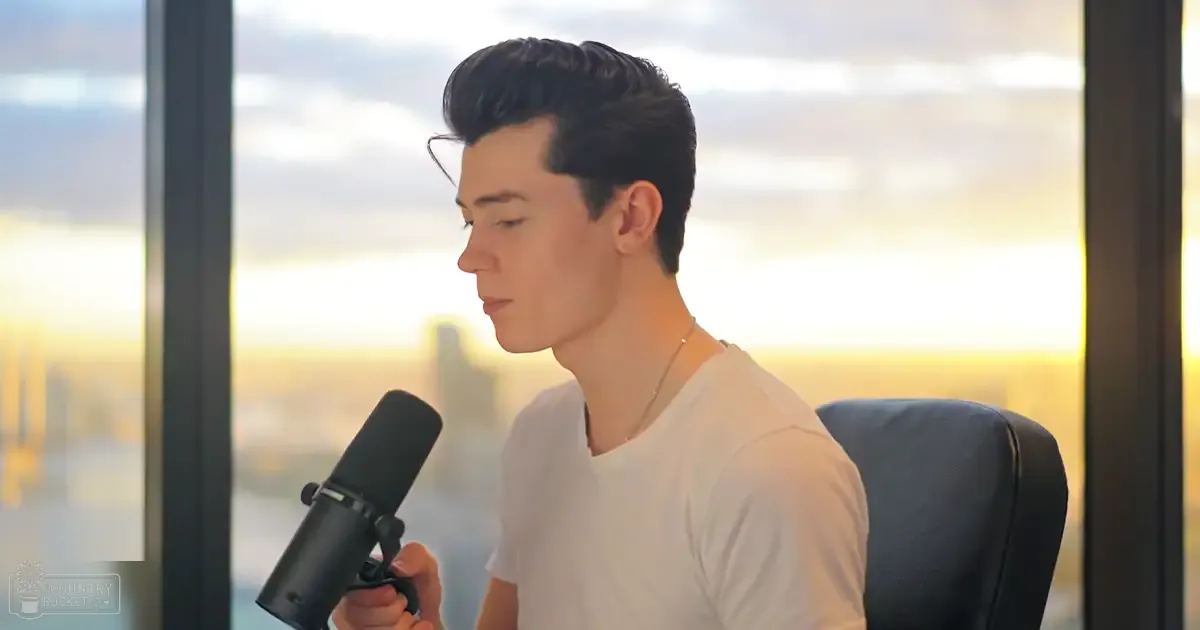 A young adult male wearing a white t-shirt with relaxed fit, round neckline, short sleeves, in an indoor setting with large windows overlooking a cityscape and sky.