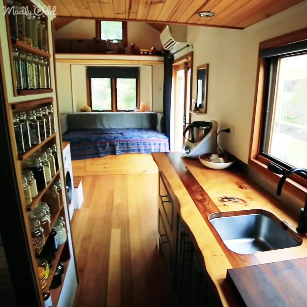 A cozy compact living space with wooden countertops, a sink, shelves filled with jars, a bed with a blue and red patterned blanket, and natural light through small windows.