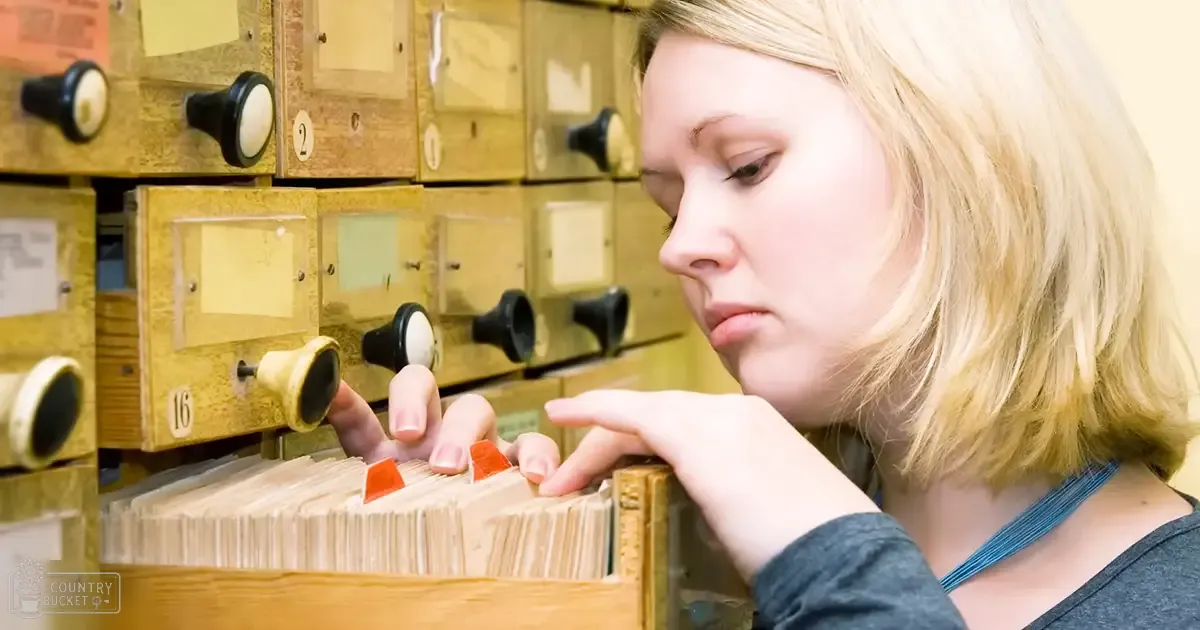 A young adult female in an archive setting wearing a dark gray long-sleeved shirt with relaxed fit and round neckline, surrounded by old-fashioned wooden card catalogs under even bright lighting.