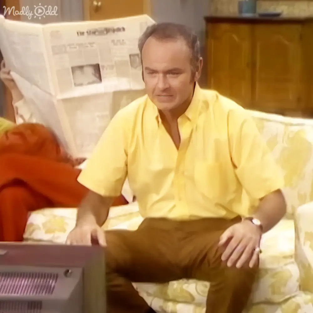 A middle-aged man wearing a yellow short-sleeved shirt and holding a wristwatch, standing indoors with a floral-patterned sofa and a vintage television in the background.