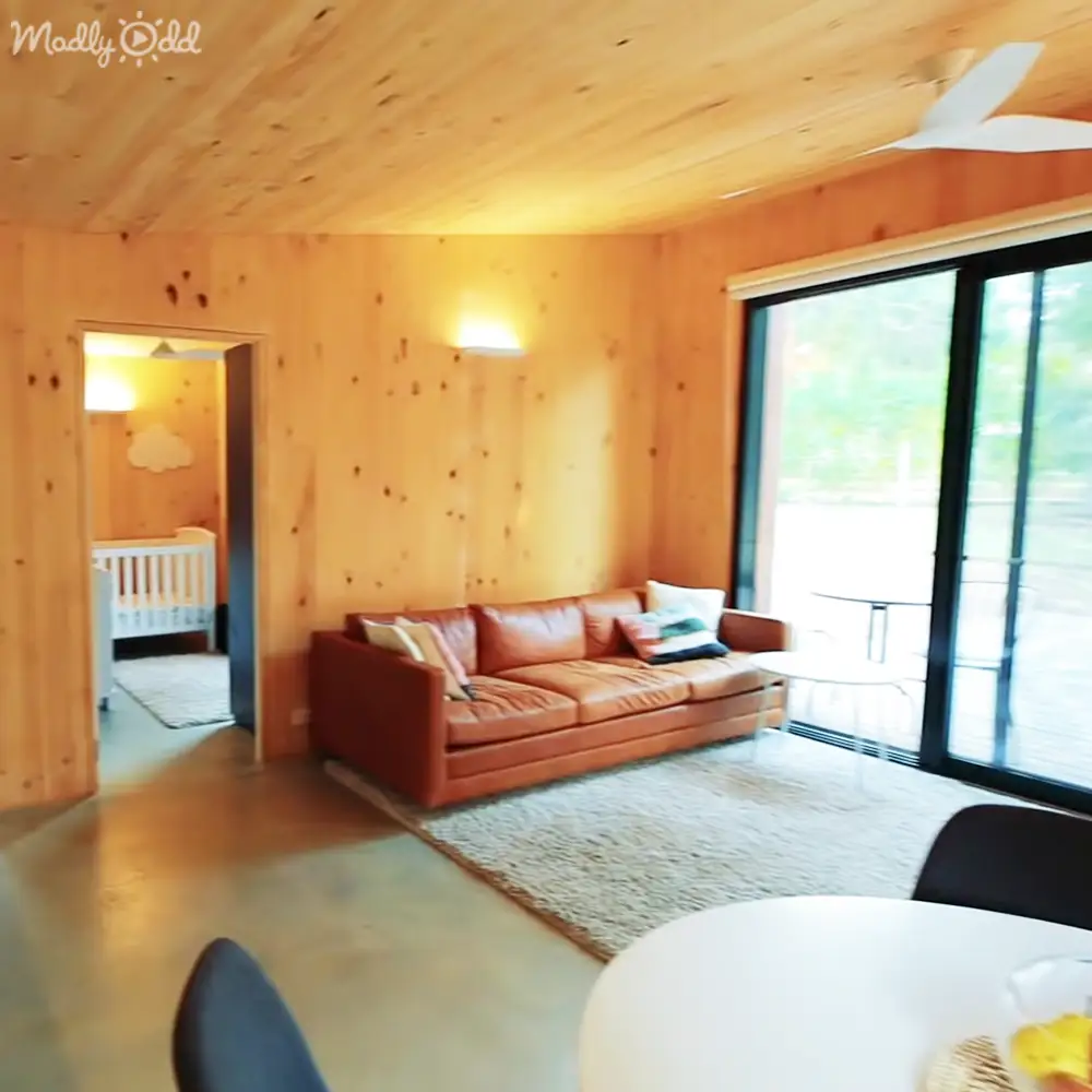 A cozy living room with wooden walls and ceiling, modern home interior with a brown leather sofa adorned with cushions, white round table, glass coffee table near sliding doors, crib in nursery area, large windows allowing natural light, greenery outside suggesting spring or summer, daytime lighting.