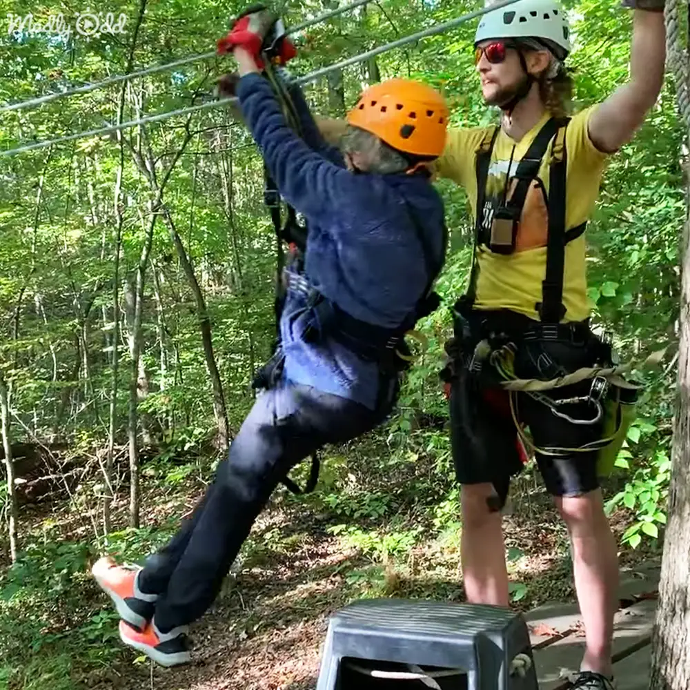 An older adult male participant wearing a blue long-sleeve shirt and orange helmet being assisted by a young adult to middle-aged male instructor in a dense forested area with green foliage, during what appears to be late morning or early afternoon sunlight filtering through the trees.