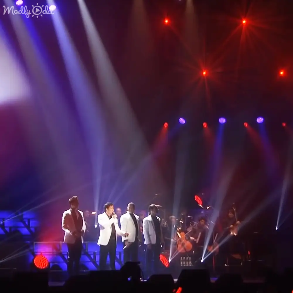 An indoor concert scene with adult male musicians in formal attire performing under vibrant red, blue, and white stage lighting at night.