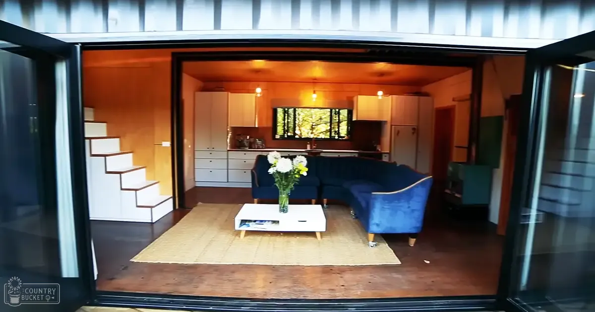Modern living space with blue sectional sofa, white coffee table, and kitchen area with white cabinets and greenery outside. Indoors during daytime.