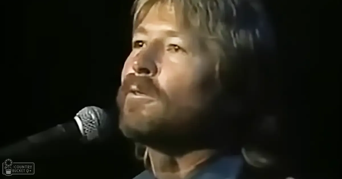 A male adult singer wearing a dark, collared shirt from the late 20th century performs into a microphone at a concert with dim stage lighting and a dark background.