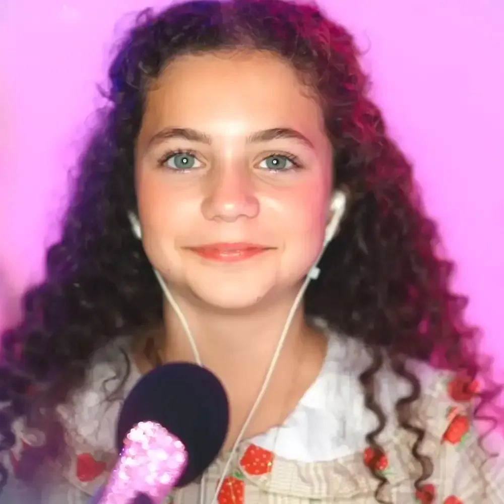 A young adult female wearing a flowy, ruffled collar white dress with red strawberry patterns holds a glittery pink microphone against a solid light purple background.