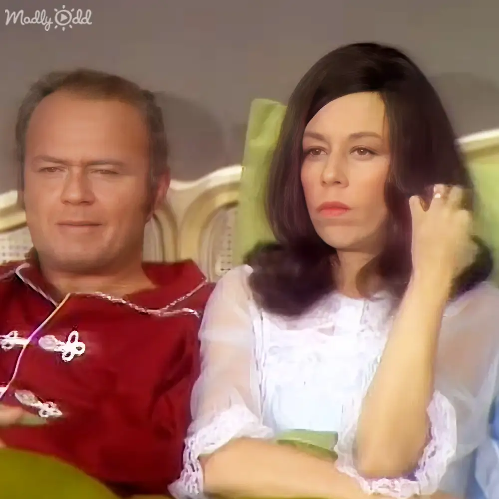 Adult woman seated on a green patterned couch with an older adult male in a red robe-like garment in a softly lit indoor living room setting.