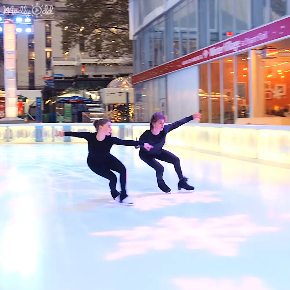 Two young female skaters in black leotards at an indoor rink within a Winter Village setting, surrounded by festive decorations under artificial evening lighting.
