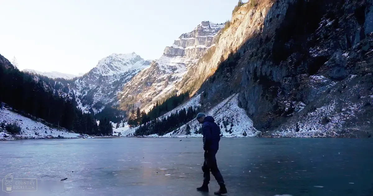 Adult male in dark blue outdoor attire, standing by a frozen lake surrounded by snow-covered mountains under a blue sky with patches of white snow.