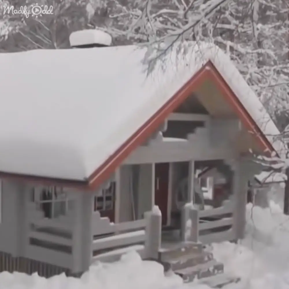 A small, snow-covered house with red trim stands amidst a winter landscape, surrounded by trees and covered in heavy snowfall.