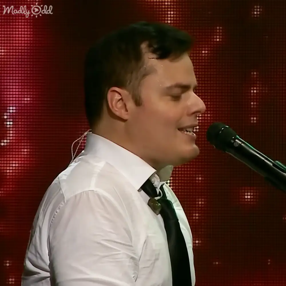 A young adult male singer wearing a white dress shirt, black tie, and headphones performs on an indoor stage with a red LED background.