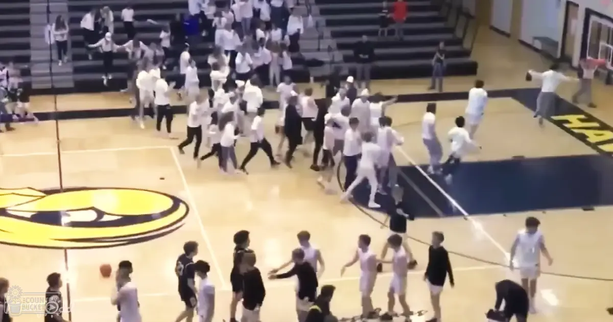 A group of young adults celebrate in a brightly lit indoor gymnasium with white t-shirts and basketball court markings.
