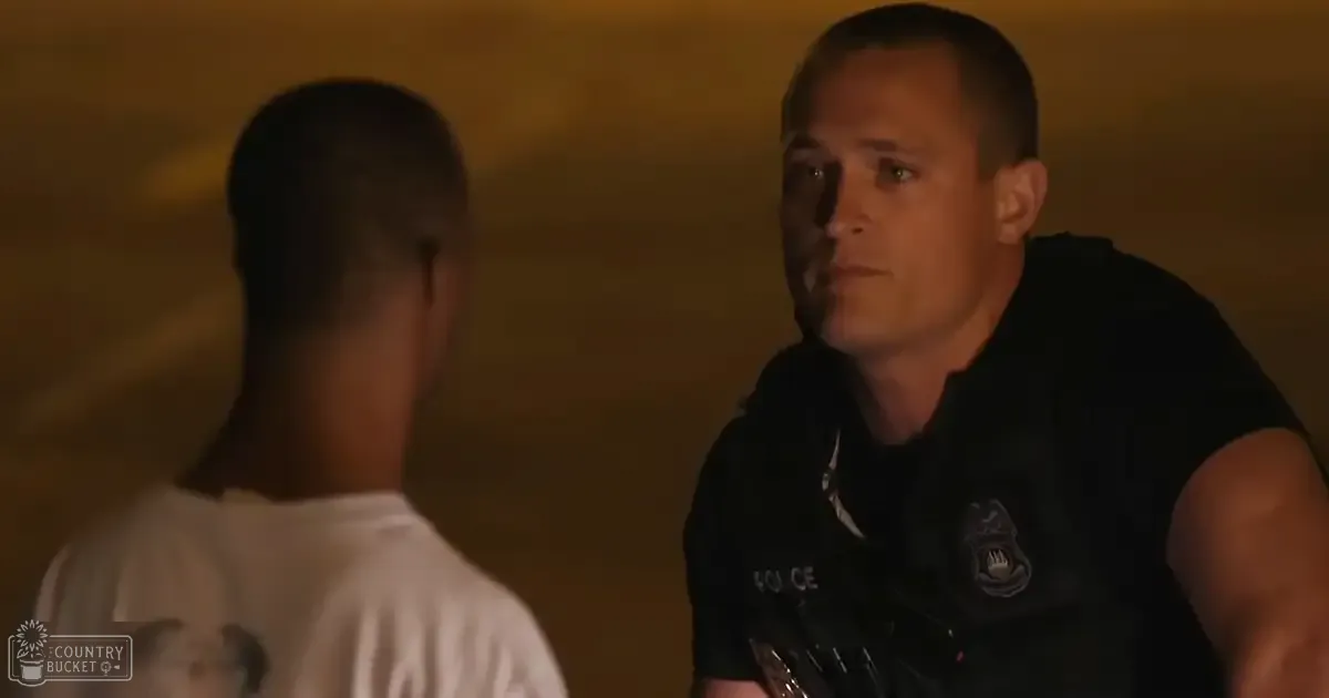 An adult male police officer and a young adult male in casual attire interact against an indoor wall with warm evening lighting.