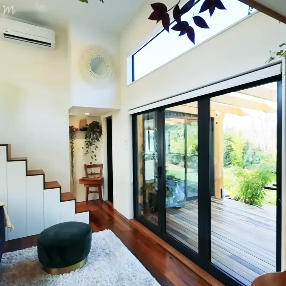 A modern interior space with a white staircase, wooden steps, a round green ottoman on a white shag rug, a wooden chair near an open doorway leading to another room, and a large sliding glass door opening onto a wooden deck.