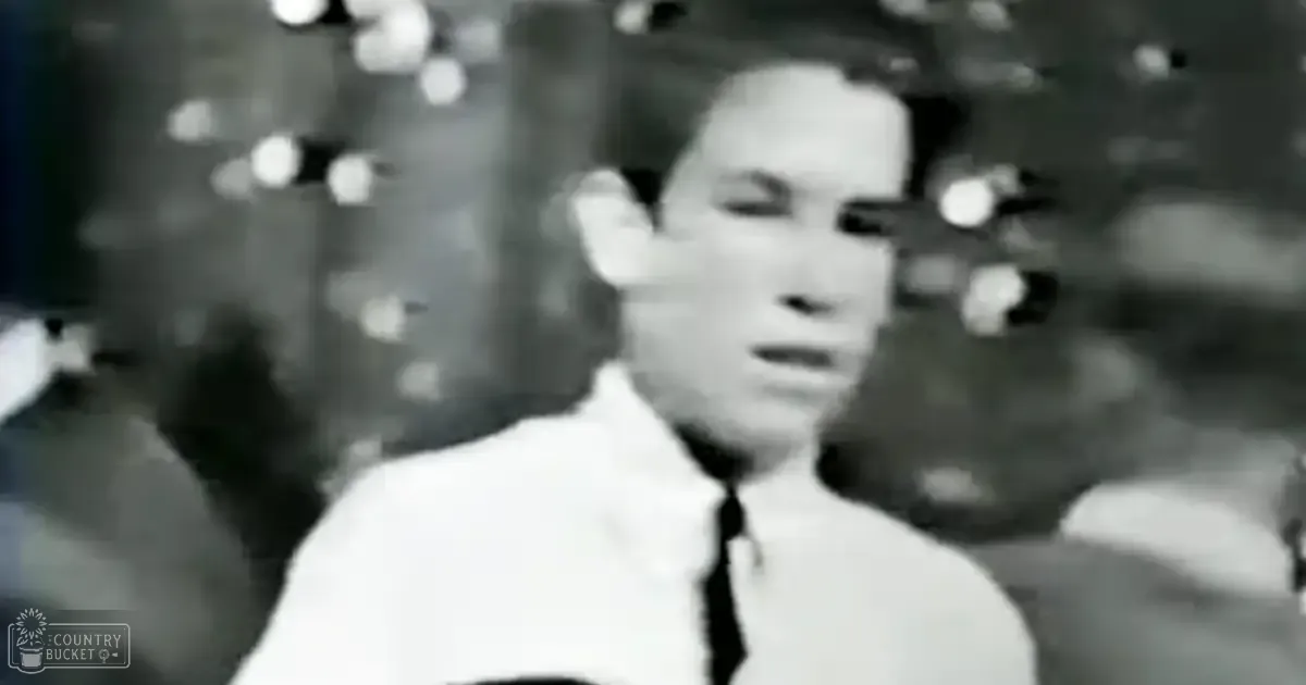 Young adult male wearing a white collared shirt and dark tie, likely from mid-20th century, in an indoor evening gathering with festive background lighting.