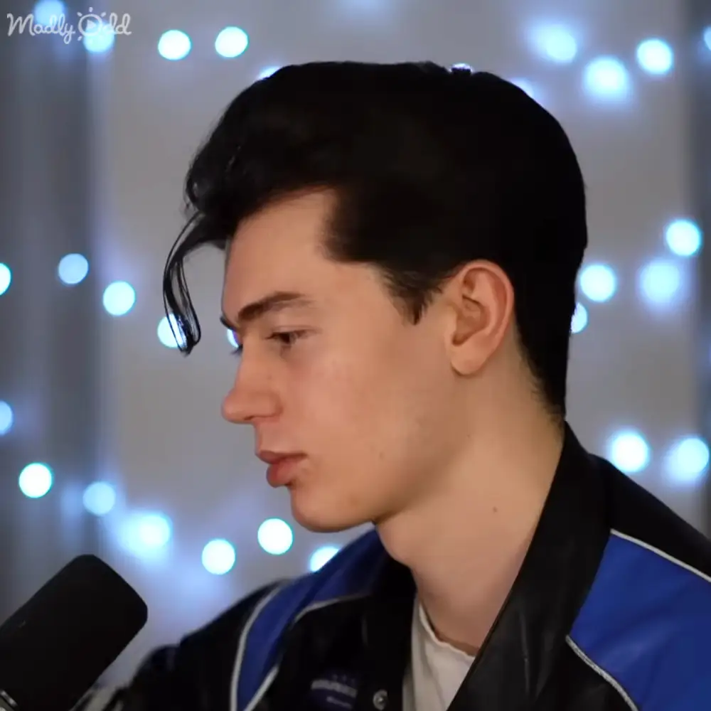 A young adult male wears a relaxed fit black jacket with blue accents and white trim, suggesting a modern casual style. The scene includes decorative fairy lights in the background and a microphone on the left side of the frame.