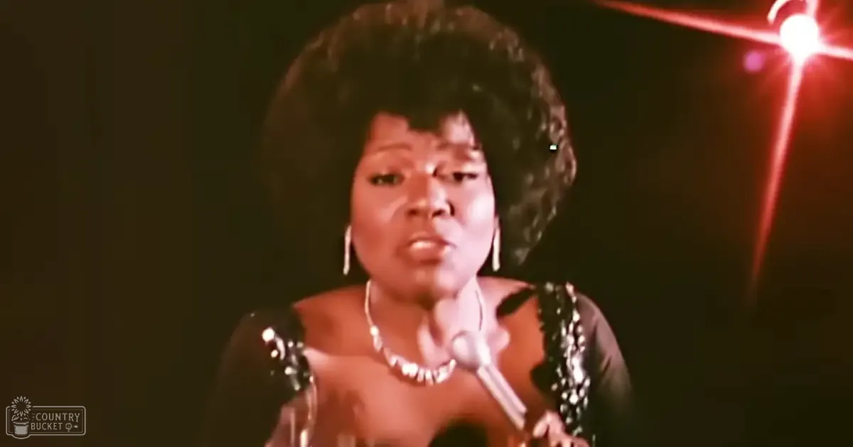 An adult female performer wearing a black, sleeveless, sequined or beaded top with large hoop earrings and a long necklace, holding a microphone on a dimly lit stage.