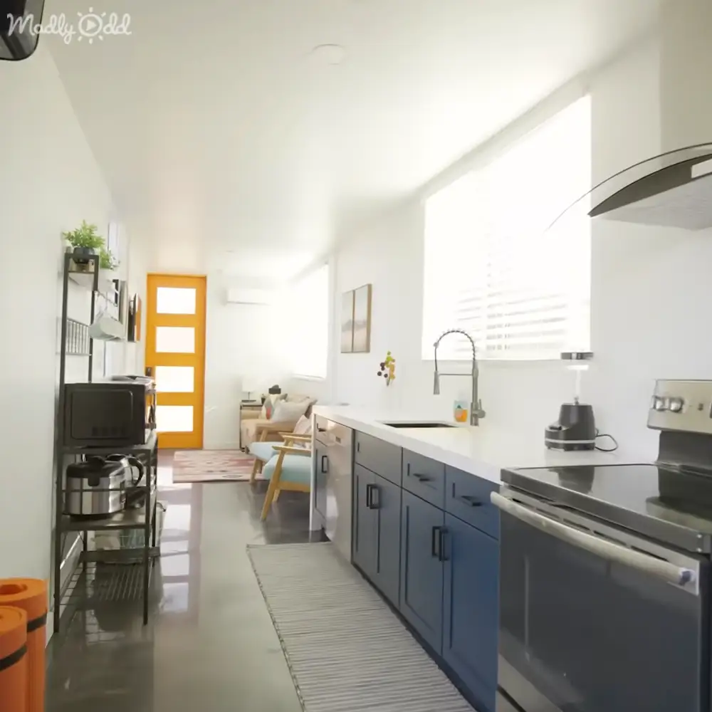 A modern kitchen with blue cabinets, a stainless steel oven, sink, countertop items, microwave on metal shelf, yellow door leading to another room, light-colored living area furniture and decor in an indoor apartment setting.