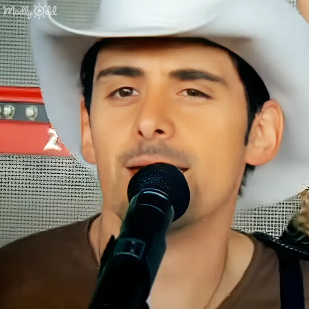 A young adult male singer wearing a fitted V-neck brown shirt, holding a microphone in an indoor setting with neutral lighting.