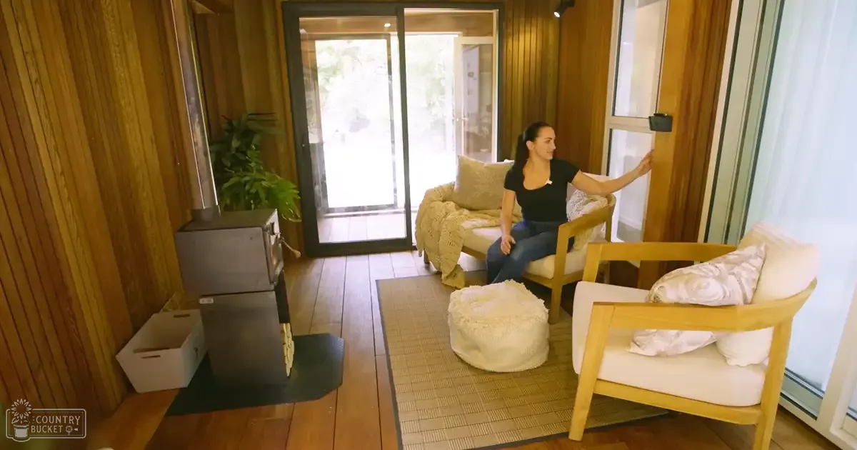 A cozy living room with wooden panel walls, sliding glass doors leading to an outdoor patio with greenery visible outside, and furniture including a wood-burning stove, two armchairs with white cushions and floral-patterned throw pillows, and a sofa covered in a beige blanket.