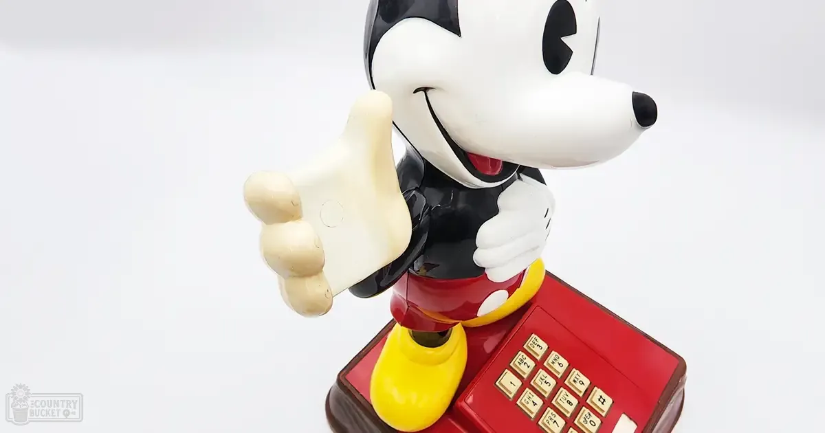 Mickey Mouse, a cartoon mouse with white face, black ears, red shorts, and yellow shoes stands on a plain white background. A classic red telephone is the focal point beneath him.